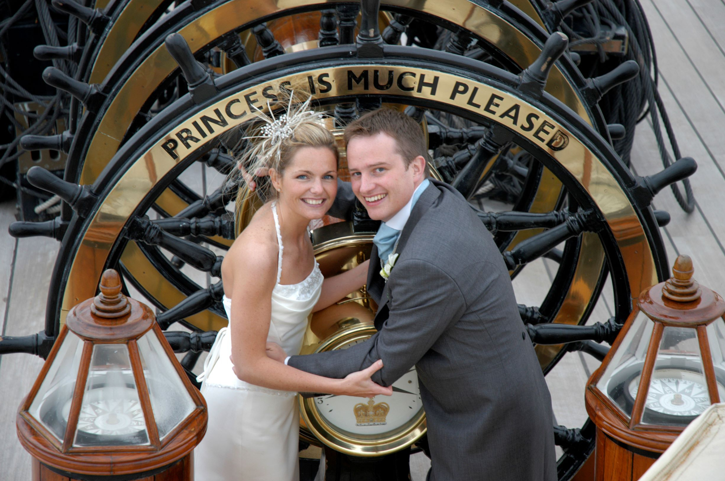 A bride and groom pose for a picture in front of HMS Warrior's ship wheel. The front wheel has a caption engraved on it that says "Princess is much pleased".