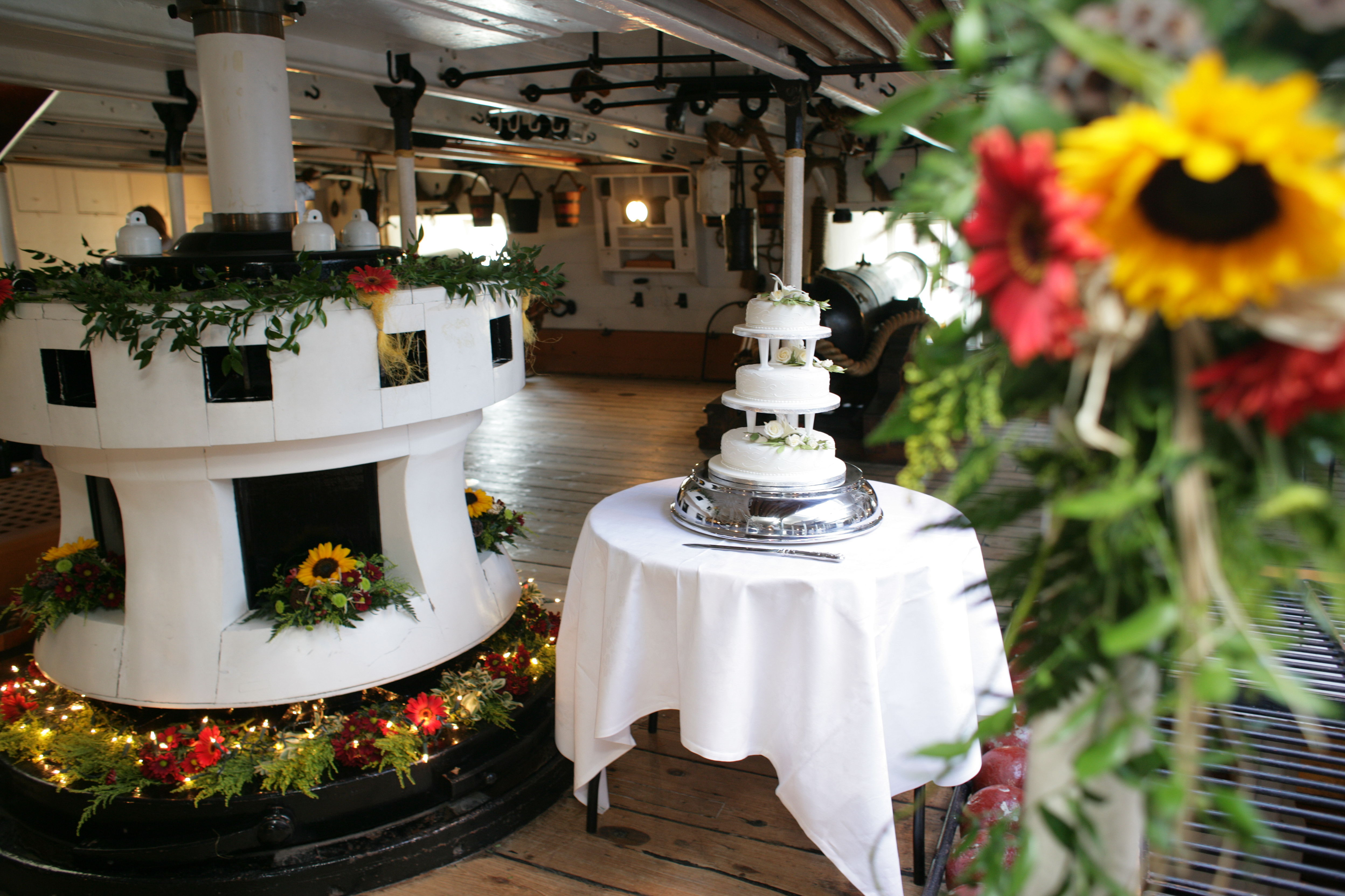 HMS Warrior's Capstan decorated with sunflowers and wildflowers for a wedding. There is a white wedding cake on a table next to it. 