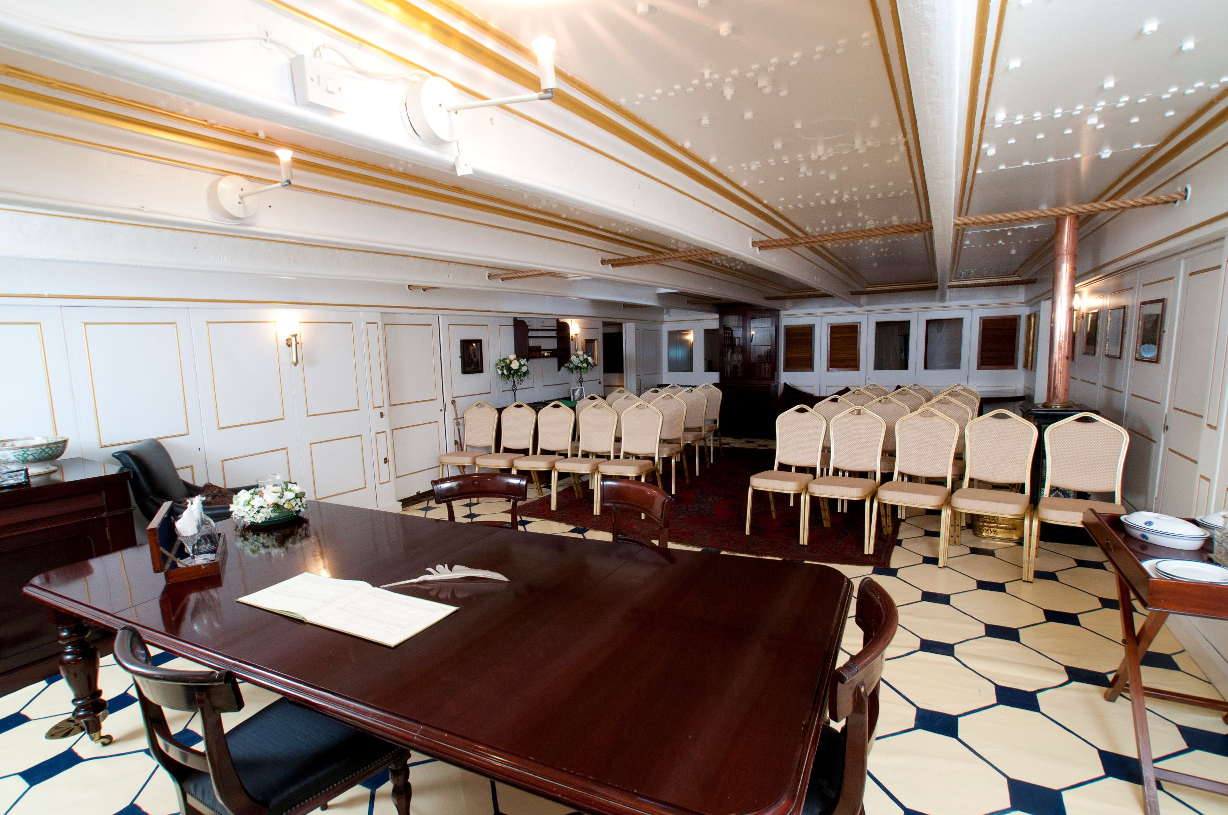 HMS Warrior's Captains Cabin set up for a wedding ceremony. The couple sit at the Captain's desk, with cream coloured seats for the guests