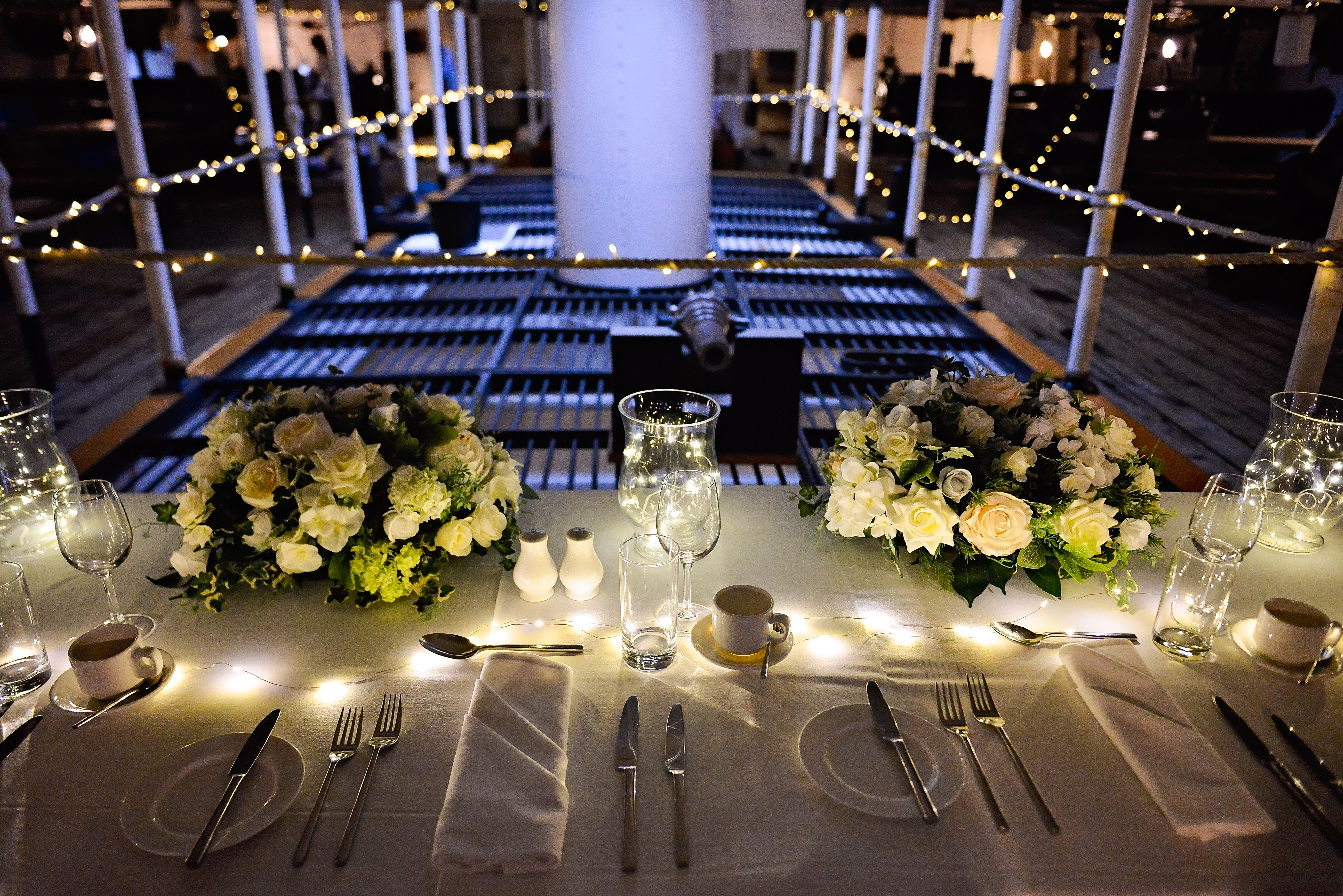 The wedding top table onboard a ceremony at HMS Warrior. The table is made up with white linen and cream flowers, and illuminated with white fairy lights. 