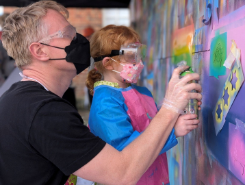 A graffiti artist shows a little girl how to use a spray can on an art wall 