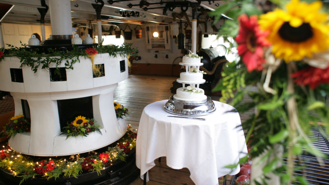 HMS Warrior's Capstan decorated with sunflowers and wildflowers for a wedding. There is a white wedding cake on a table next to it. 