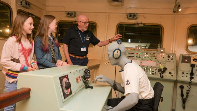 A volunteer show two young girls what the bridge of an aircraft carrier looks like